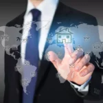 Businessman in suit touches virtual house symbol on world map - symbolic image for international property investment and asset management.