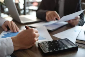 Two businessmen analyse financial documents with laptop, calculator and diagrams on a desk