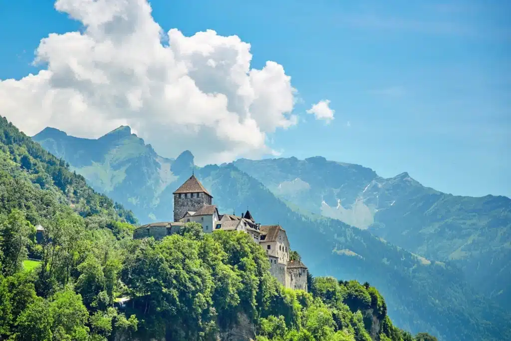Das Schloss Vaduz auf einem grünen Hügel, Berge und Wolken zieren den Himmel der Fürstentum Liechtenstein Stiftung.