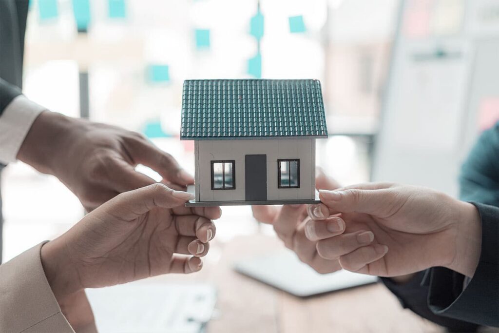 Drei Personen, die ein kleines Modellhaus zusammenhalten, als Symbol für Teamwork oder Zusammenarbeit im Immobilienbereich.