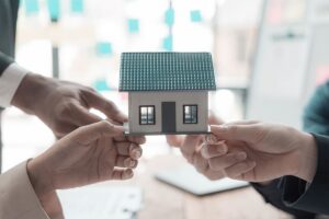 Drei Personen, die ein kleines Modellhaus zusammenhalten, als Symbol für Teamwork oder Zusammenarbeit im Immobilienbereich.